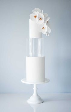 a three tiered cake with white flowers on top is displayed in front of a gray background