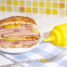 a ham and cheese sandwich on a plate next to a fork, knife and mustard bottle