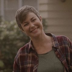 a woman with short hair smiling and wearing a flannel shirt in front of a house