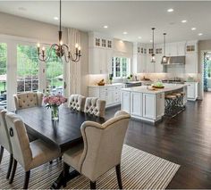 a dining room table and chairs in front of an open kitchen area with large windows