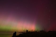 two people are standing on the shore watching the aurora lights in the sky over water