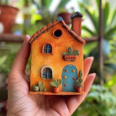 a hand holding a miniature house made out of clay with plants in the back ground