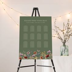 an easel with a seating chart on it next to a vase and string lights