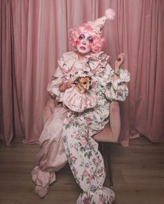 a woman with pink hair sitting in a chair holding a small dog and wearing a clown mask