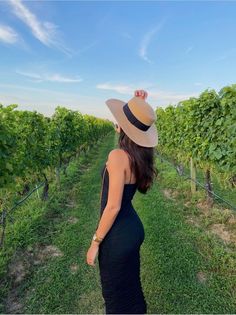 a pregnant woman in a black dress and straw hat walks through a vineyard with green vines