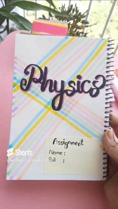 a hand holding a spiral notebook with the word physics written in black on it