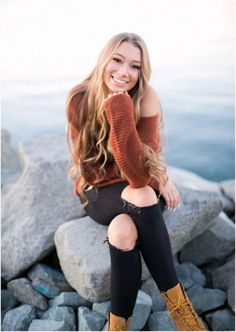 a beautiful young woman sitting on top of a rock next to the ocean wearing boots