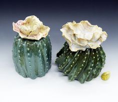 two ceramic flowers sitting on top of each other next to a green cactus plant with yellow seeds