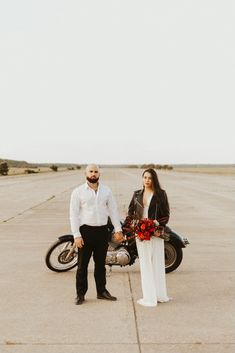 a man and woman standing next to a motorcycle in the middle of an empty parking lot