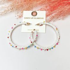 Pictured are raffia braided hoop earrings in white with colorful beads. They are pictured with a pink feather on a white background. White Beaded Earrings For Spring Party, Trendy Hoop Earrings For Vacation, Handmade White Trendy Beaded Earrings, White Handmade Trendy Beaded Earrings, Handmade Trendy White Beaded Earrings, Handmade White Beaded Earrings Trendy Style, Bohemian White Beaded Earrings For Spring, White Hoop Earrings For Summer, White Bohemian Hoop Earrings With Colorful Beads