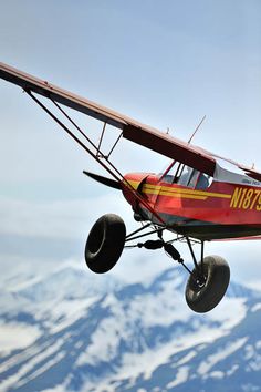 a small red airplane flying in the air with mountains in the background