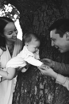 a man and woman holding a baby next to a tree with their hands on the child's chest