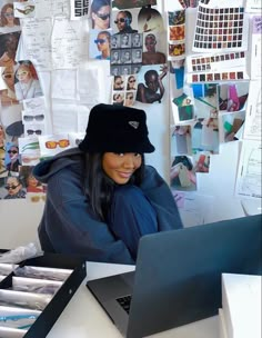 a woman sitting at a table with a laptop computer in front of her and lots of pictures on the wall behind her