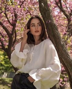 a woman standing next to a tree with pink flowers on it's branches and wearing a white blouse