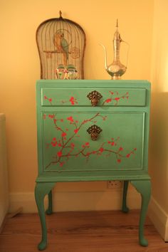 a green dresser with red flowers painted on the front and side drawers, next to a birdcage