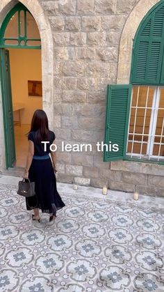 a woman in a black dress is walking towards a building with green shutters and the words to learn this