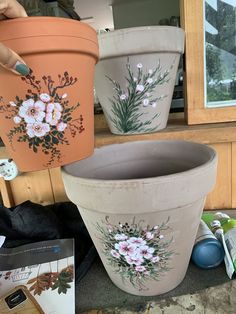 three flower pots with flowers painted on them sitting on the ground next to other potted plants