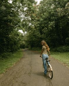 a woman riding a bike down a dirt road in the middle of some trees and grass