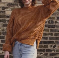 a woman wearing a brown sweater and jeans standing in front of a brick wall with her hand on her head