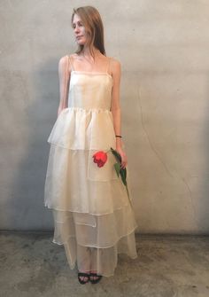 a woman in a white dress holding a flower and standing against a wall with her hands on her hips