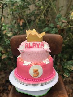 a pink and red cake with a crown on top sitting on a chair in front of some bushes