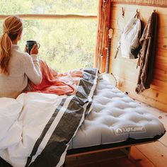 a woman sitting on top of a bed next to a window holding a coffee cup