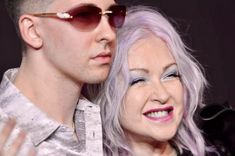 an older woman with pink hair and a shaved head next to a young man wearing sunglasses