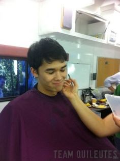 a young man getting his hair cut by another person in the background with other people around him