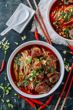 two bowls of beef noodle soup with chopsticks and red peppers on the side