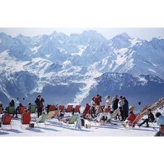 a group of people sitting in lawn chairs on top of a snow covered slope with mountains in the background