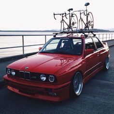 a red car with two bikes on top of it's roof rack and parked next to the water