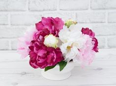 a white vase filled with pink and white flowers on top of a wooden table next to a brick wall