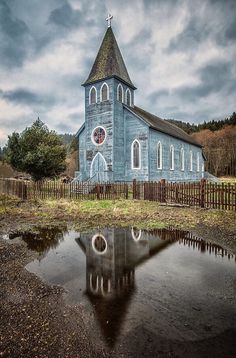 an old blue church with a steeple and stained glass windows is reflected in the water