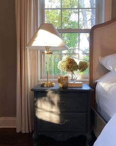 a nightstand with flowers on it next to a bed in front of a large window