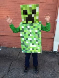 a young boy in a minecraft creeper costume standing on the street with his hands up