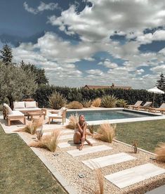 a woman sitting in front of a pool surrounded by grass and rocks with lounge chairs around it