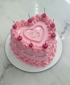 a pink heart shaped cake with cherries on top sits on a white plate next to a marble countertop