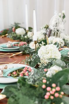 the table is set with green and white flowers, greenery, candles and plates