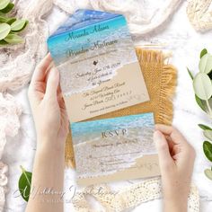 a person holding up two wedding cards in front of some flowers and greenery on the beach