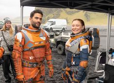 two people in orange spacesuits standing next to each other near cars and trucks