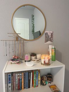 a white book shelf with books and candles on it next to a large round mirror