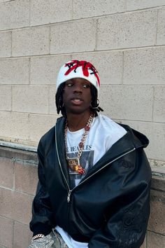 a young man sitting against a brick wall wearing a jacket and hat with red bows on it