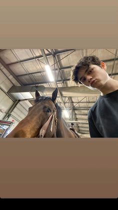 a young man standing next to a brown horse in a warehouse with other horses behind him