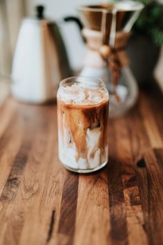 a glass filled with liquid sitting on top of a wooden table next to two coffee pots