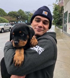 a man holding a puppy in his arms on the sidewalk with cars parked behind him