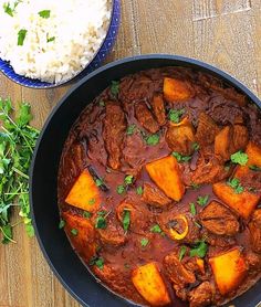 a pan filled with stew and rice on top of a wooden table next to a bowl of rice