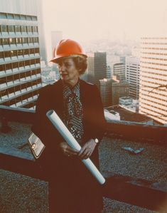 an older woman wearing a hard hat and holding a large piece of paper on top of a building