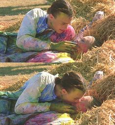 two men laying in hay with their faces covered by colored paint and looking at cell phones