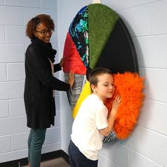 a woman standing next to a young boy in front of a wall hanging with different colored feathers on it