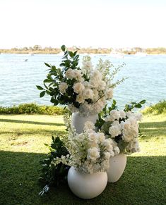 three white vases with flowers in them sitting on the grass near some water and trees